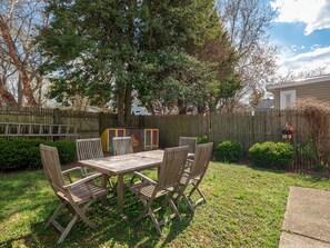 Un ensemble de table et de chaises en bois, installé dans un jardin, avec une clôture en bois, un arbre et une maison en arrière-plan.