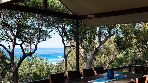 Una terraza de madera con mesa y sillas, con vistas a la costa, árboles y el mar.