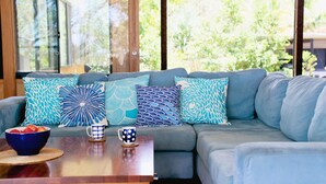 Una sala de estar moderna con un sofá gris decorado con cojines estampados, una mesa de centro de madera con un cuenco de fruta y dos tazas, y ventanas amplias que ofrecen vistas a zonas verdes.