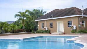 Ein Schwimmbecken mit gefliestem Rand, ein Haus mit braunem Dach und Palmen im Hintergrund.