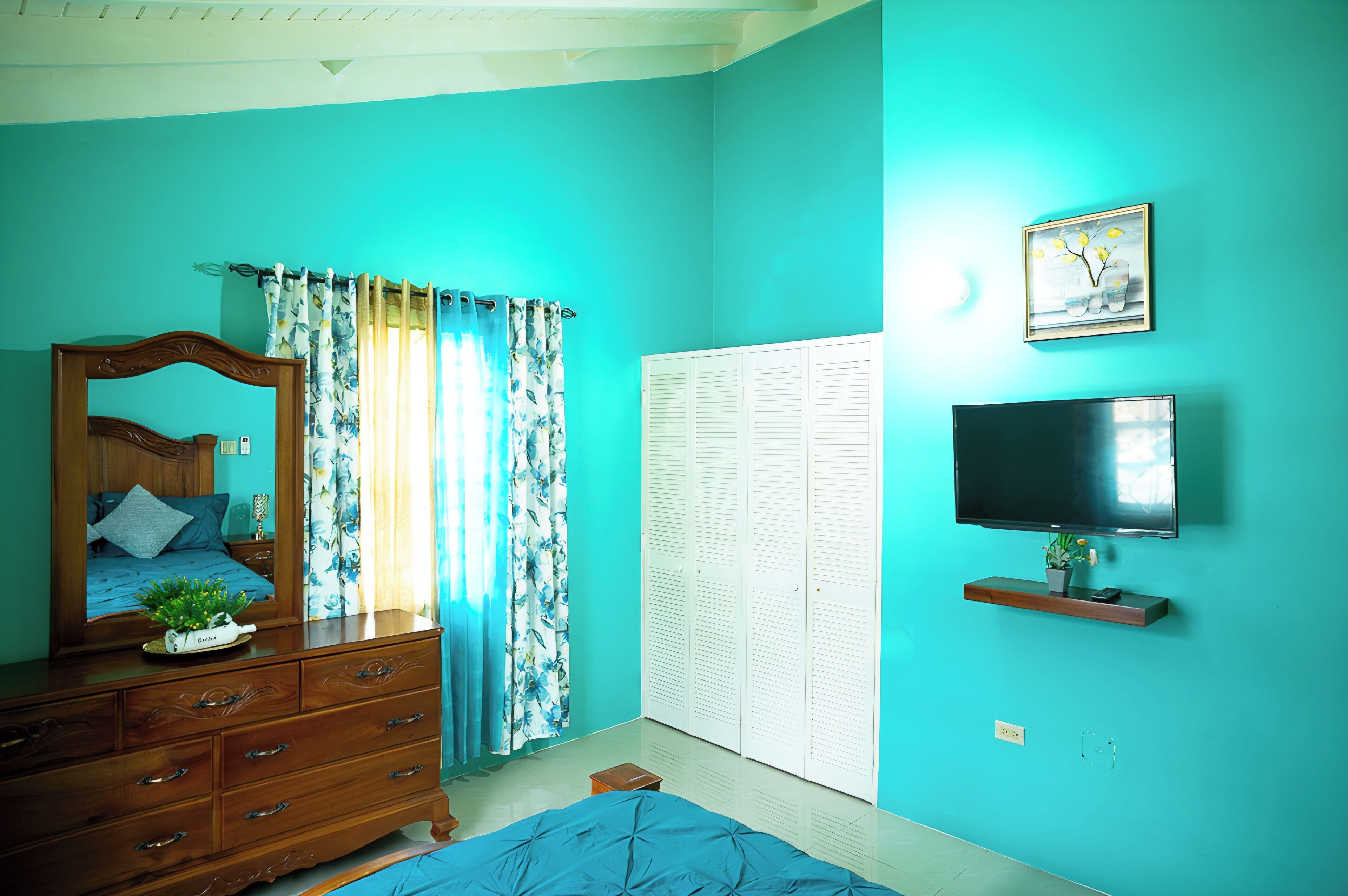 A bedroom with teal walls, a wooden dresser, a bed with blue bedding, a television on a shelf, and a window with floral curtains.
