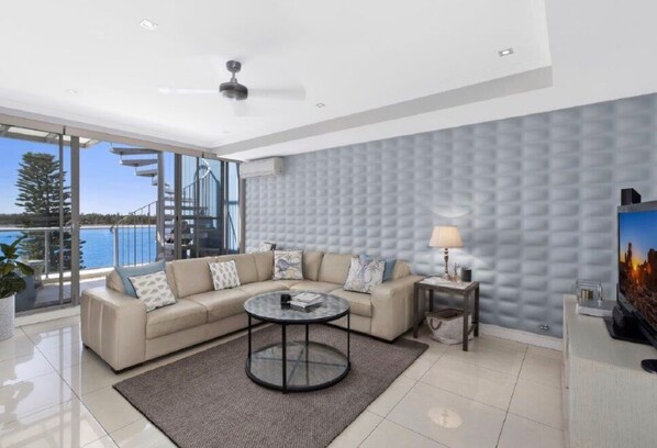 Una sala de estar moderna con sofá, mesa de centro y televisor, que cuenta con un ventanal con vistas al mar y un balcón.