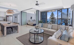 Una sala de estar moderna con sofá, mesa de centro y comedor. Hay un ventanal con vista al mar.
