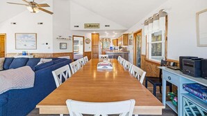A spacious living room with a wooden dining table, blue sofa, and white chairs. A ceiling fan and a kitchen area are visible in the background. - One Grand Whim | 698 ft from the Beach | Community Pool, Hot Tub | Corolla (Corolla)