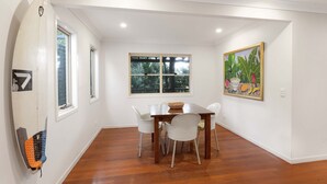 A dining area with a wooden table and chairs, a surfboard, and a window with a view of greenery. - Spacious home with views suitable for 2 families (Point Lookout)
