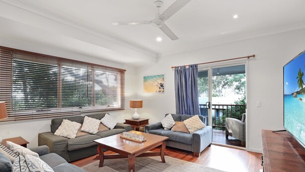 A spacious living room with a ceiling fan, two sofas with patterned cushions, a wooden coffee table, and a view of a balcony with outdoor furniture. - Spacious home with views suitable for 2 families (Point Lookout)
