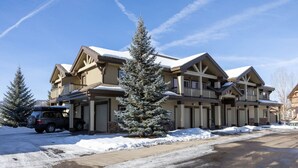 Un chalet à étages, dont le toit est recouvert de neige, avec une allée couverte et un VUS garé devant.