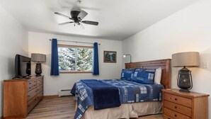 Une chambre à coucher comprenant un lit, une commode en bois, des tables de chevet, un ventilateur de plafond et une fenêtre avec des rideaux.
