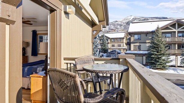 Un balcon agrémenté de fauteuils en osier et d’une petite table, offrant une vue sur un paysage montagneux enneigé et sur d’autres bâtiments.