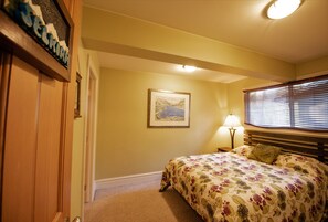 Un dormitorio con cama, mesita de noche, lámpara, un cuadro enmarcado en la pared y una puerta.