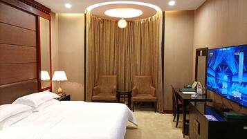 A hotel room with a bed, two armchairs, a desk with a television, and a large window with curtains.