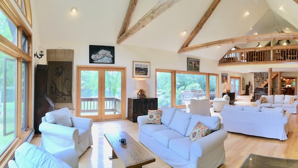 A spacious living area with multiple sofas, a wooden coffee table, and large windows offering a view of the outdoors. - Spruce Lake Farm 56-acre mountain-top log home estate on private 7-acre lake (Bovina)
