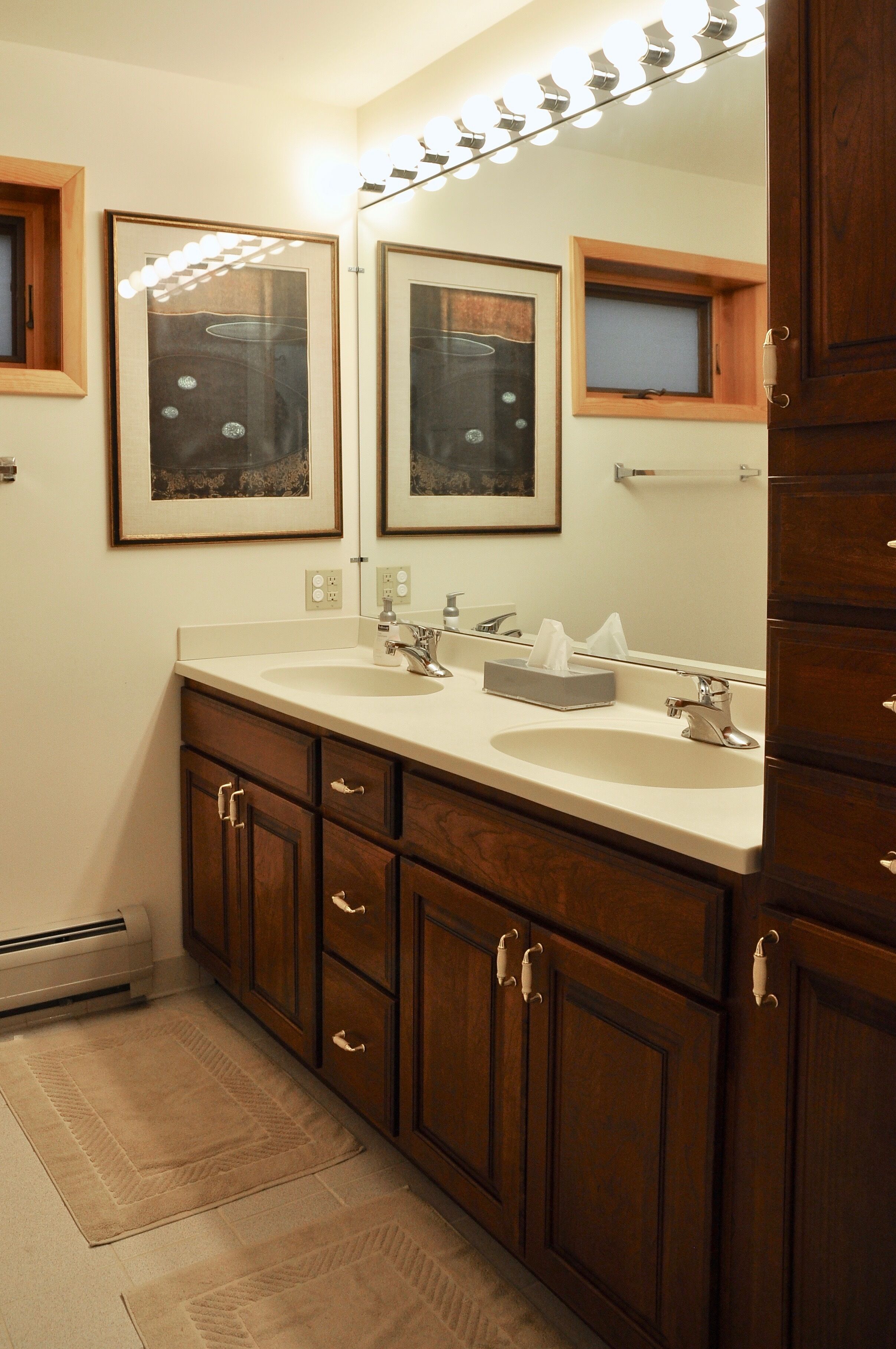 A bathroom with a double sink, wooden cabinets, a large mirror, and framed artwork on the wall.
