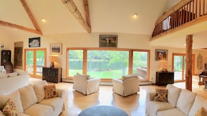 A spacious living room with multiple seating areas, large windows offering views of the outdoors, a grand piano, and a staircase leading to an upper level. - Spruce Lake Farm 56-acre mountain-top log home estate on private 7-acre lake (Bovina)