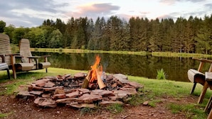 A campfire surrounded by rocks, with Adirondack chairs and a lake in the background. - Spruce Lake Farm 56-acre mountain-top log home estate on private 7-acre lake (Bovina)