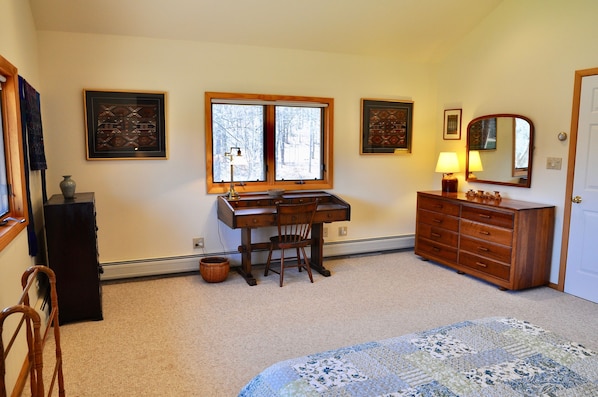 A bedroom with a desk, chair, dresser, and a large window.