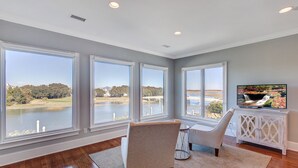 A spacious living room with large windows overlooking a lake, a TV on a white cabinet, and a cozy seating area. - 20 Morgan Place Drive by AvantStay | Waterfront Home w/ Private Dock (Isle of Palms)