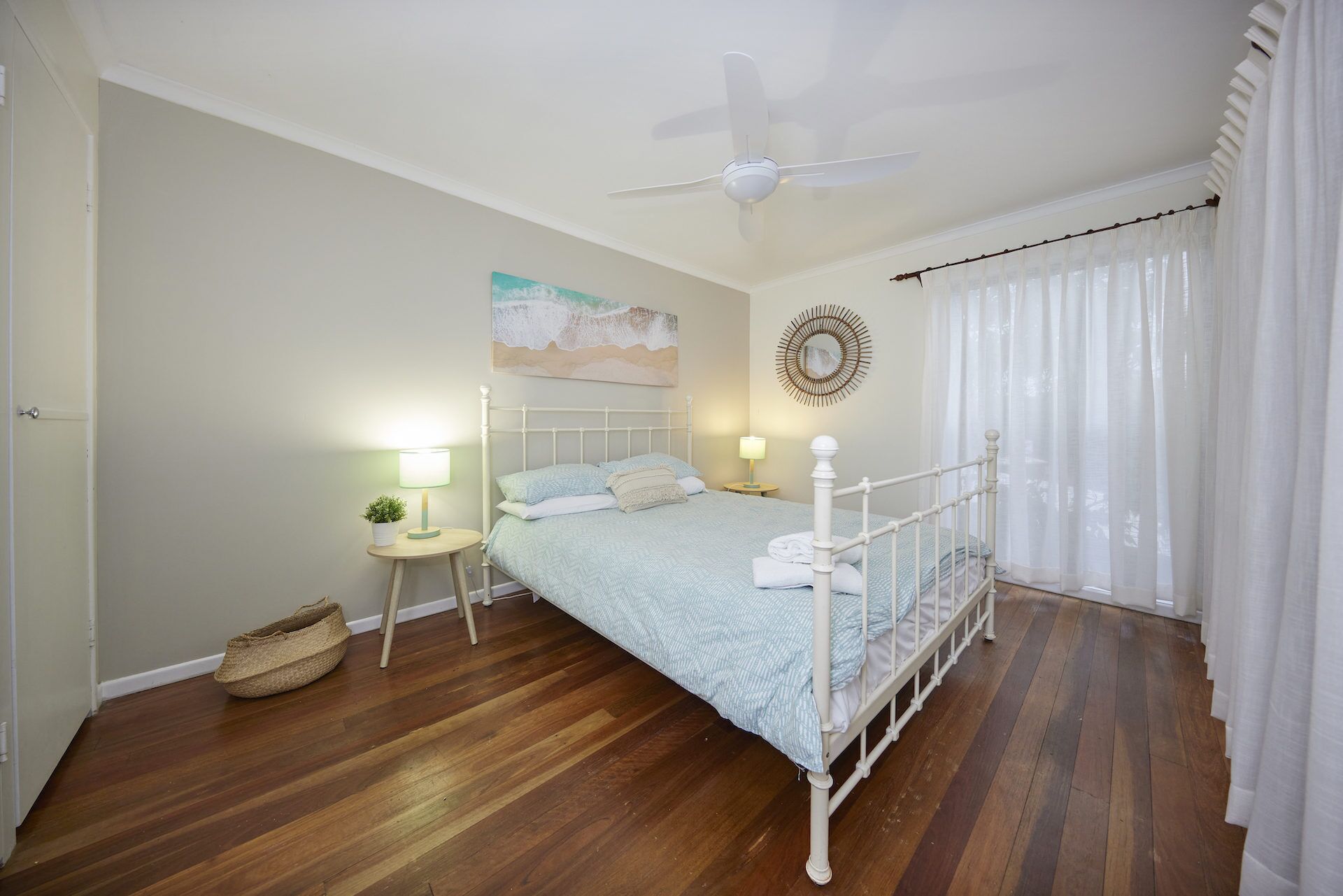 A bedroom with a bed, a ceiling fan, a bedside table with a lamp, a wall decoration, and a window with curtains.