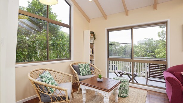 Une pièce chaleureuse, avec des fauteuils en osier, une table en bois et une bibliothèque. De grandes fenêtres offrent une vue sur la verdure environnante.