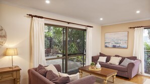 Un salon avec un canapé, une table basse et une lampe. Il y a une fenêtre avec des rideaux, un tableau au mur et une vue sur la verdure à l’extérieur.