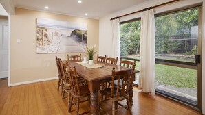 Un coin repas avec une table et des chaises en bois, un tableau représentant une plage et une grande fenêtre avec des rideaux.