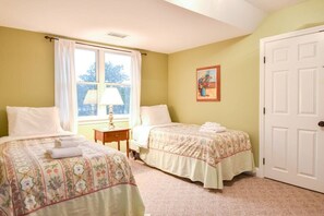 A bedroom with two beds, a window with curtains, a bedside table with a lamp, and a door.