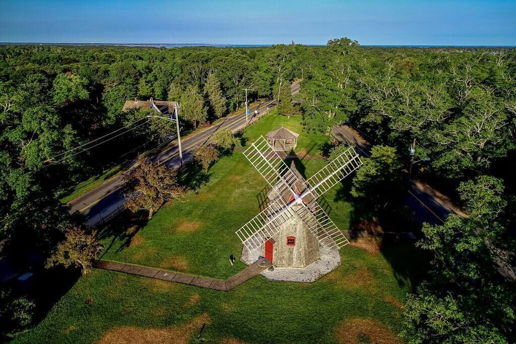 Eine Windmühle, umgeben von einer Wiese, mit einer Straße und Bäumen im Hintergrund.