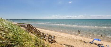 A beach with a wooden walkway leading to the water, a sandy area with a blue canopy, and a clear sky.