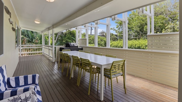 A covered outdoor patio with a white table and yellow chairs, surrounded by white railings and large windows offering a view of greenery.