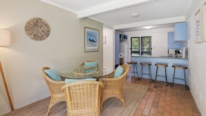 A dining area with wicker chairs, a glass table, and a view into a kitchen with a refrigerator and bar stools.