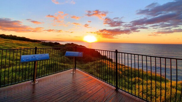 Ein Sonnenuntergangsblick von einer Holzdielenterrasse mit Metallgeländer, mit Blick auf eine Rasenfläche und das Meer.