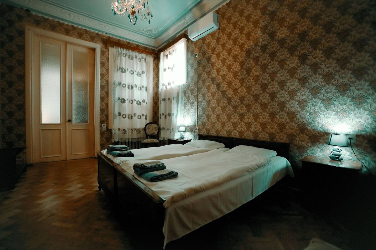 A bedroom with two beds, a chandelier, an air conditioner, and patterned wallpaper.