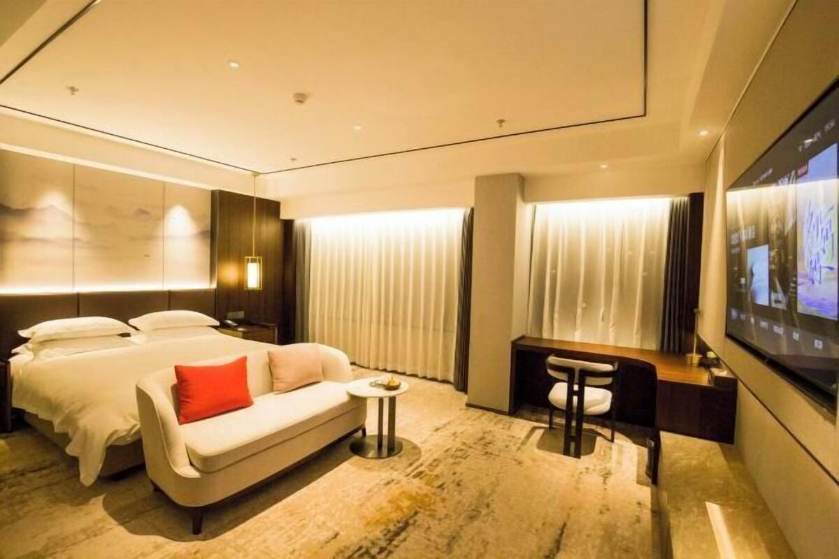 A hotel room with a bed, a sofa, a small table, a desk, a TV, and a large window with curtains.