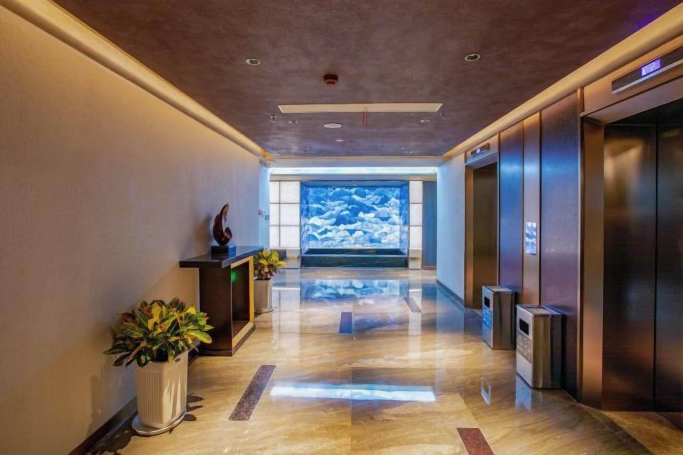 A modern hotel lobby with a polished floor, reception desk, potted plants, and elevators.