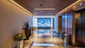 A modern hotel lobby with a polished floor, reception desk, potted plants, and elevators.