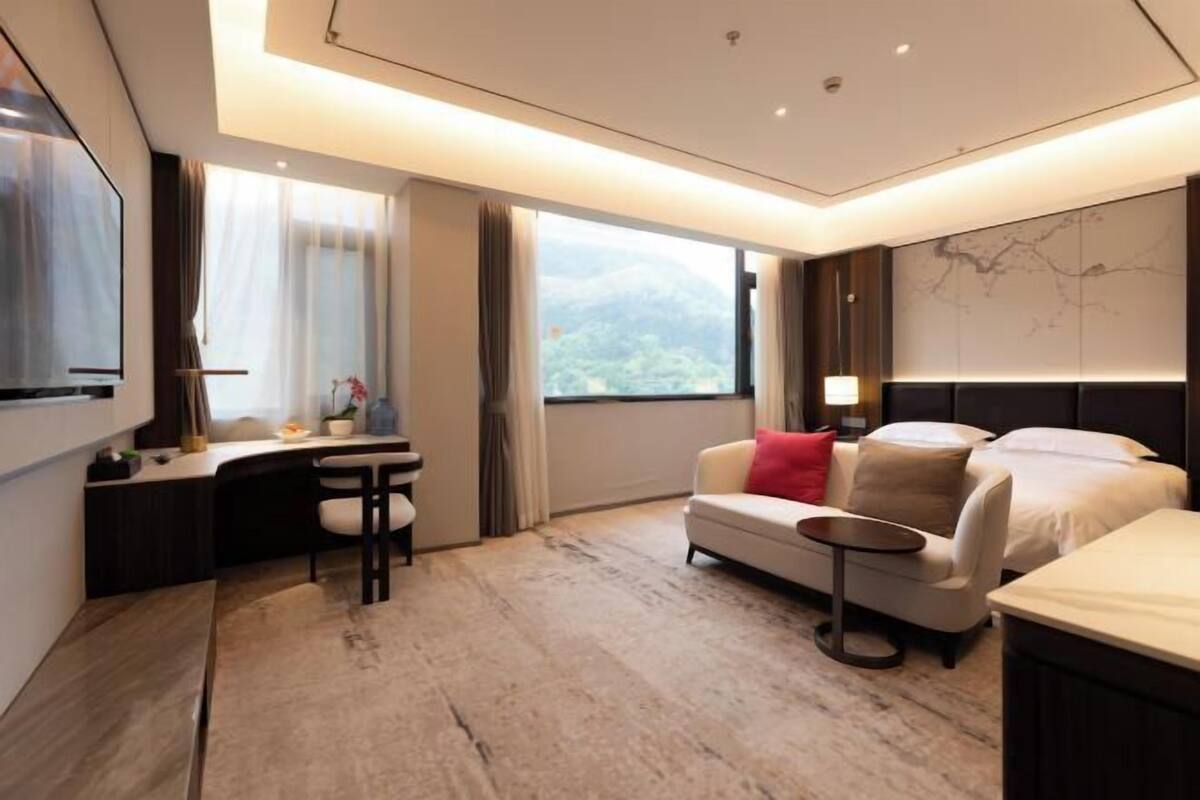A modern hotel room with a desk, sofa, and bed, large windows with a mountain view, and a flat-screen TV.