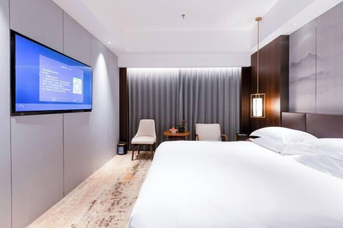 A modern hotel room with a large bed, a flat-screen TV mounted on the wall, a small table with a lamp, and a view of the city through the window.