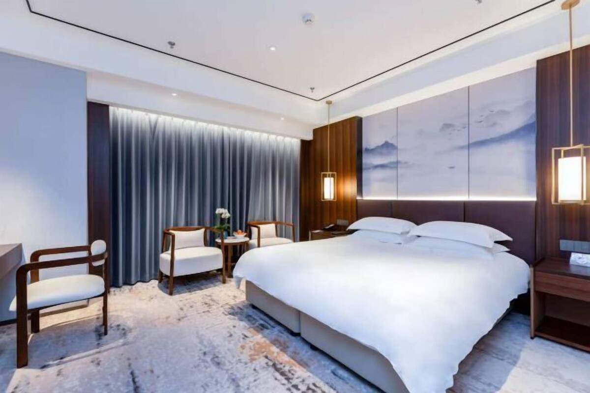 A modern hotel room with a large bed, two armchairs, a small table with a vase, and a wall-mounted TV.