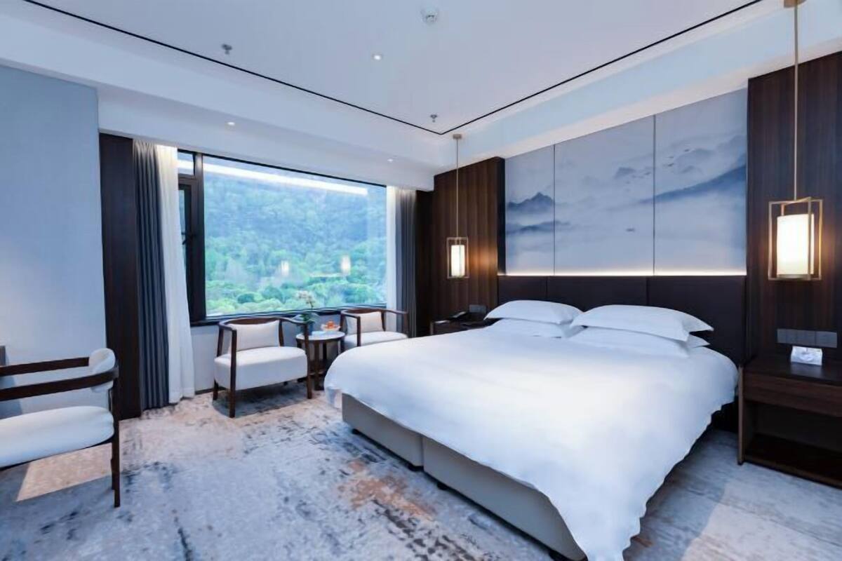 A modern hotel room with a large bed, a desk, a chair, and a view of a mountainous landscape.