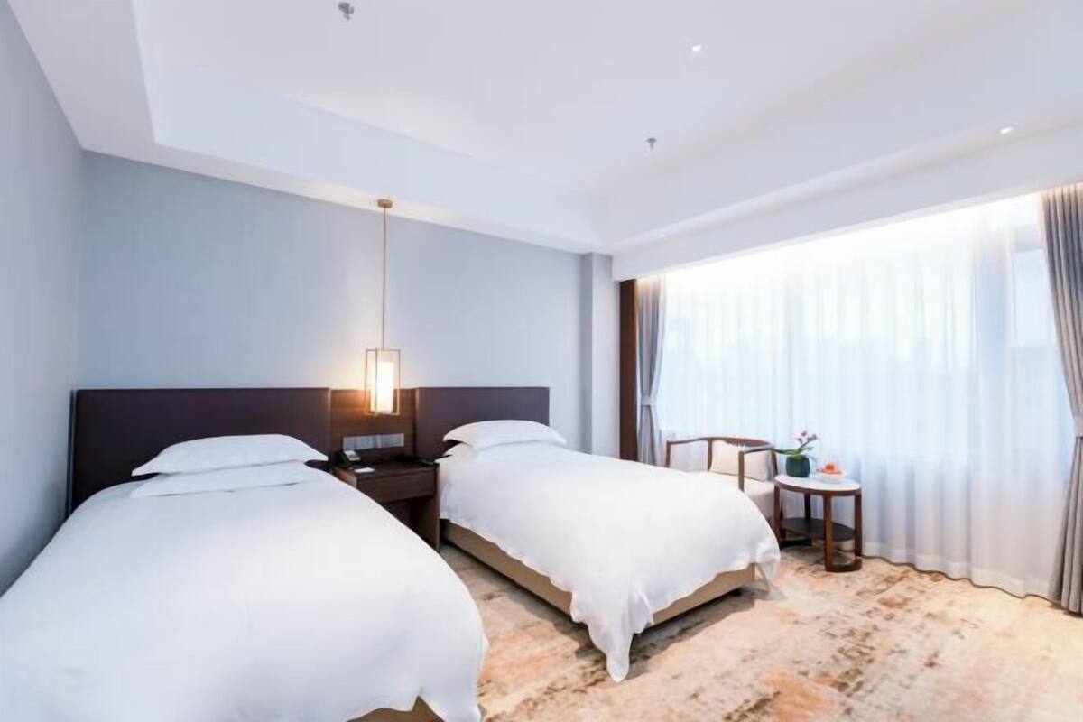 A hotel room with two beds, a nightstand, a lamp, a small table with a vase, and a large window with curtains.