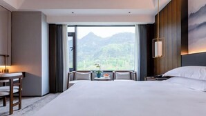 Quarto de hotel com uma cama grande, uma escrivaninha com cadeiras, vista para montanhas e uma mesa de jantar com um vaso.