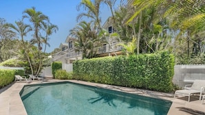 A swimming pool with a tiled deck, surrounded by palm trees and a hedge.