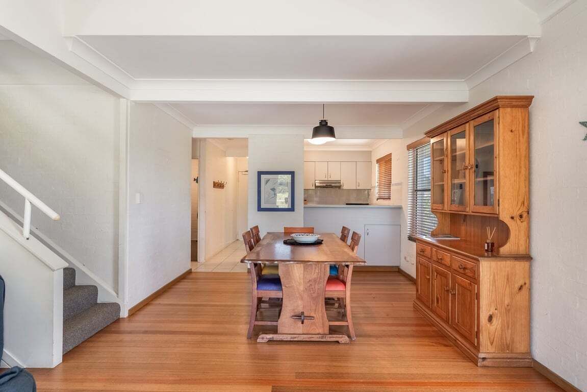 Un comedor con mesa y sillas de madera, un mueble con puertas de vidrio y una escalera a la izquierda.