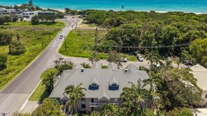 An aerial view of a coastal residential area with a prominent building featuring a flat roof and multiple chimneys, surrounded by lush greenery and a clear blue ocean in the background.