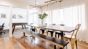 Un'ampia sala da pranzo con un lungo tavolo in legno, sedie nere e una panca. Sono presenti due lampadari a più luci, un'ampia finestra con tende leggere e quadri appesi alle pareti.
