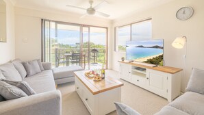 A modern living room with a grey sofa, a wooden coffee table with snacks and drinks, a TV on a wooden stand, and a balcony with a view of the sea. - Intrepid, 12/3 Intrepid Close - right on the beachfront (Nelson Bay)