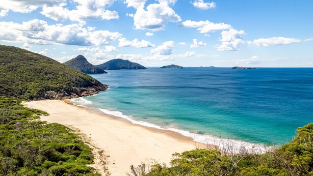 A coastal landscape with a sandy beach, clear turquoise waters, and lush green hills. - Intrepid, 12/3 Intrepid Close - right on the beachfront (Nelson Bay)