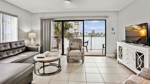 A modern living room with a sofa, armchair, and TV, a balcony with a view of palm trees and buildings, and a dining area with a table and chairs. - Land's End 5-201 - Beautifully Updated! (Treasure Island)