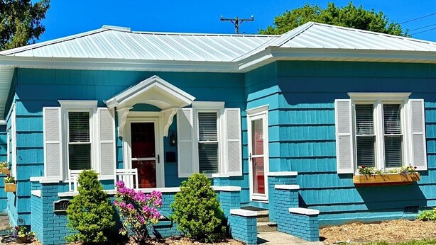 A single-story house with a gabled roof, a front porch with a white railing, and a front door with a red door frame. - Adorable, “ Beach View Cottage” Lake Michigan views! Best location!  (Ludington)