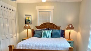 A bedroom with a wooden bed, two bedside tables with lamps, a small wooden stool, a checkered rug, a ceiling fan, and a window with blinds. - Adorable, “ Beach View Cottage” Lake Michigan views! Best location!  (Ludington)